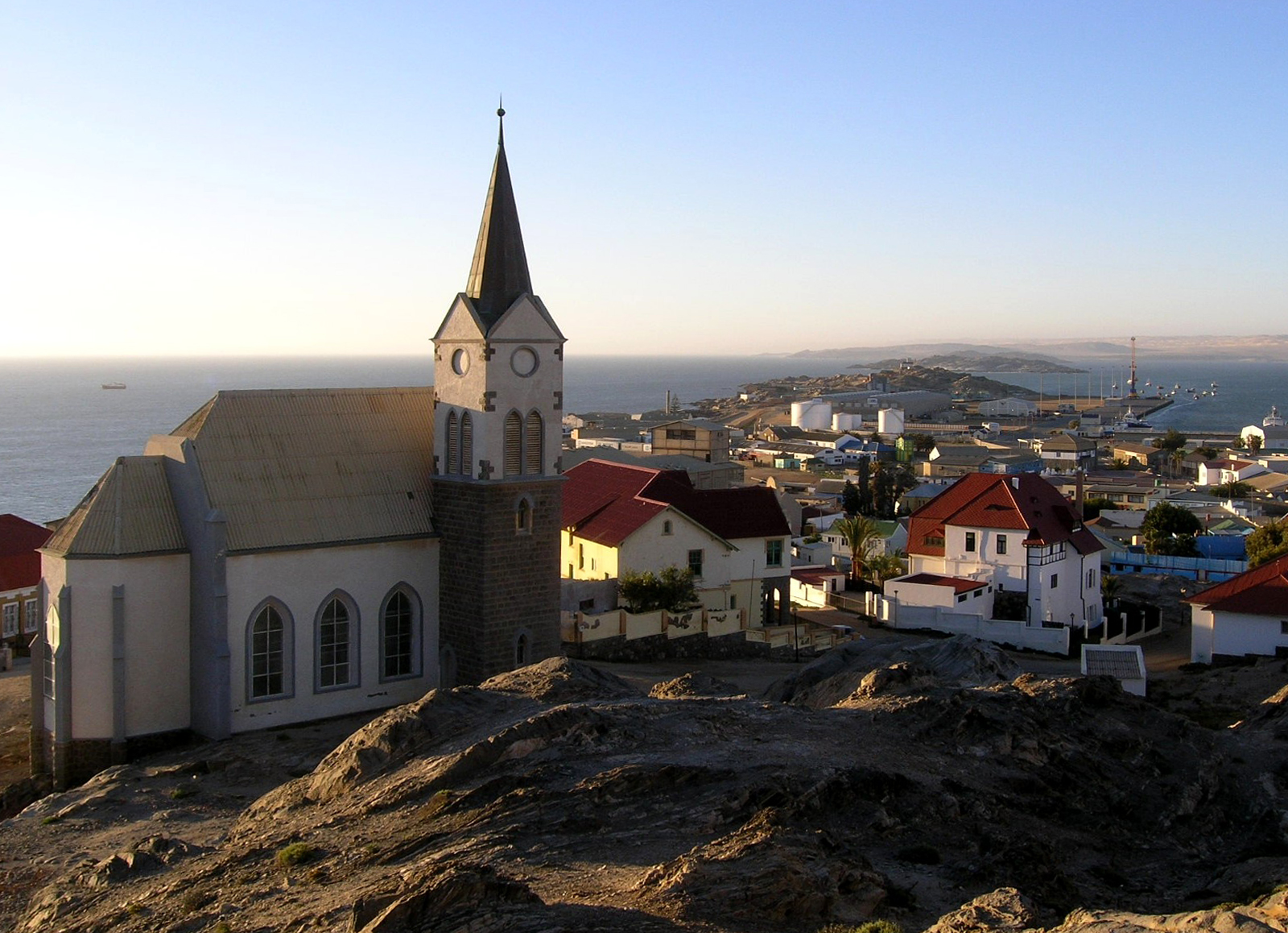 Blick auf die Felsenkirche in der Hafenstadt Lüderitz (Dr. Matthias Donath) Im Vordergrund steht eine Kirche mit spitzem Turm und großen Fenstern auf felsigem Untergrund. Die Kirche ist von hellen und roten Dächern umgeben. Im Hintergrund erstreckt sich eine Küstenstadt bis zum Meer, das im Abendlicht liegt. Die Häuser sind überwiegend niedrig gebaut und das Licht wirft lange Schatten. Am Horizont ist die Küstenlinie mit weiteren Gebäuden und einem Hafenbereich zu erkennen.