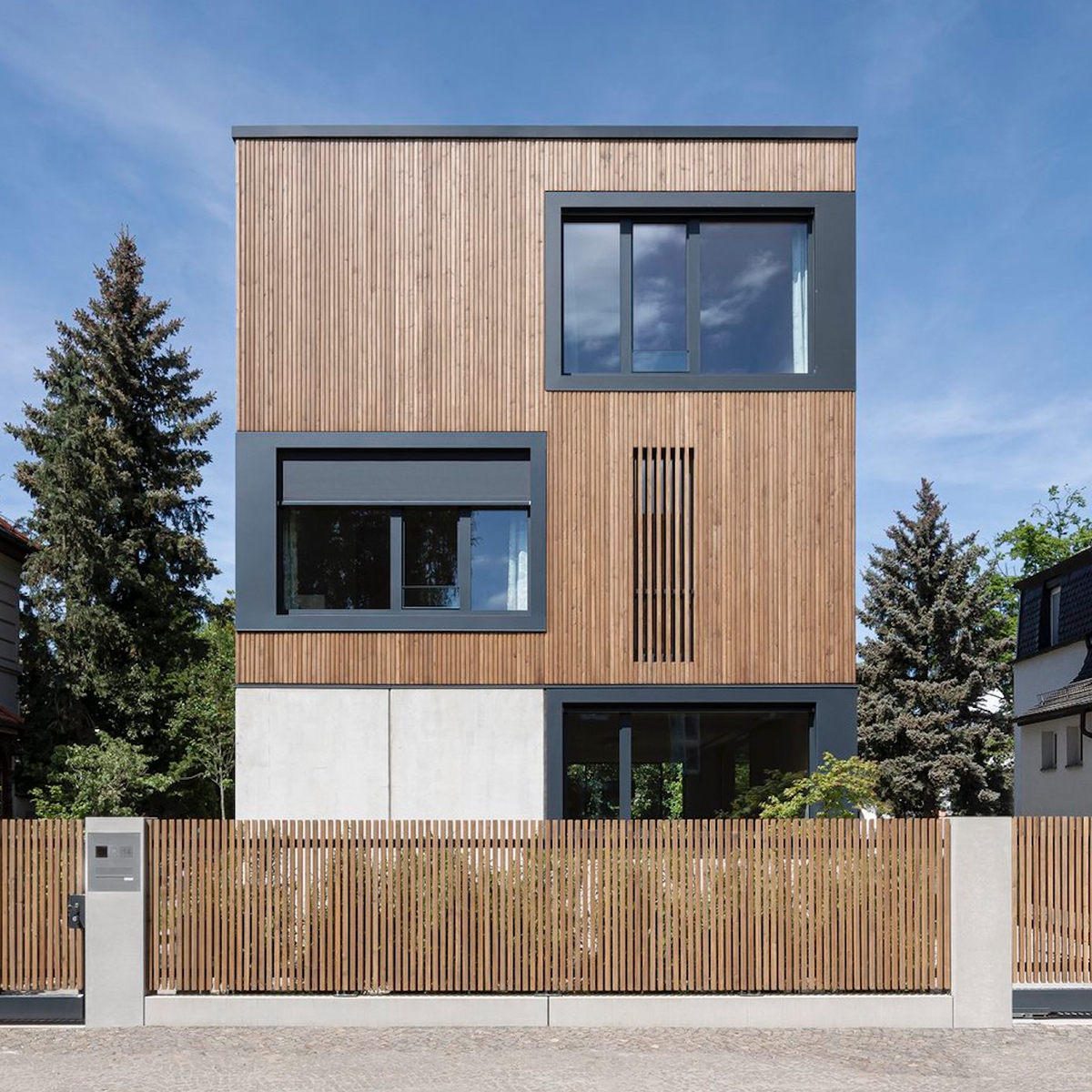 Modernes dreistöckiges Haus mit Holzfassade, großen rechteckigen Fenstern und Holzlattenzaun im Vordergrund