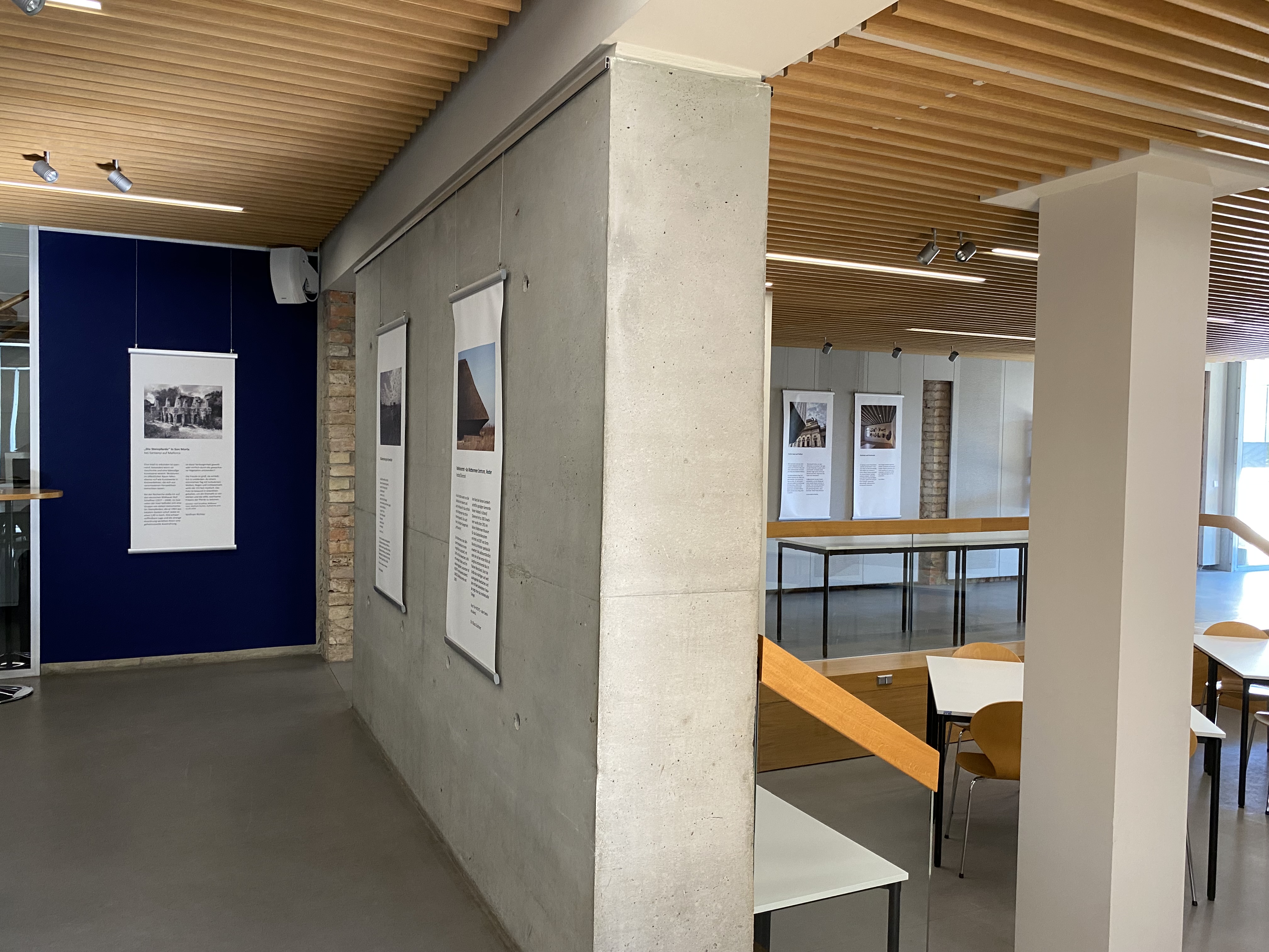 Ein Ausstellungsraum mit modernen Elementen wie einer Holzlamellendecke und Sichtbetonwänden. An den Wänden hängen mehrere gerahmte Tafeln mit Texten und Fotografien. Im Vordergrund stehen Tische und Stühle mit hellen Holzflächen. Die Beleuchtung erfolgt durch eingelassene Deckenstrahler. Im Hintergrund ist ein Geländer aus Holz und Glas zu sehen, das einen offenen Bereich abgrenzt, in dem Tische stehen.