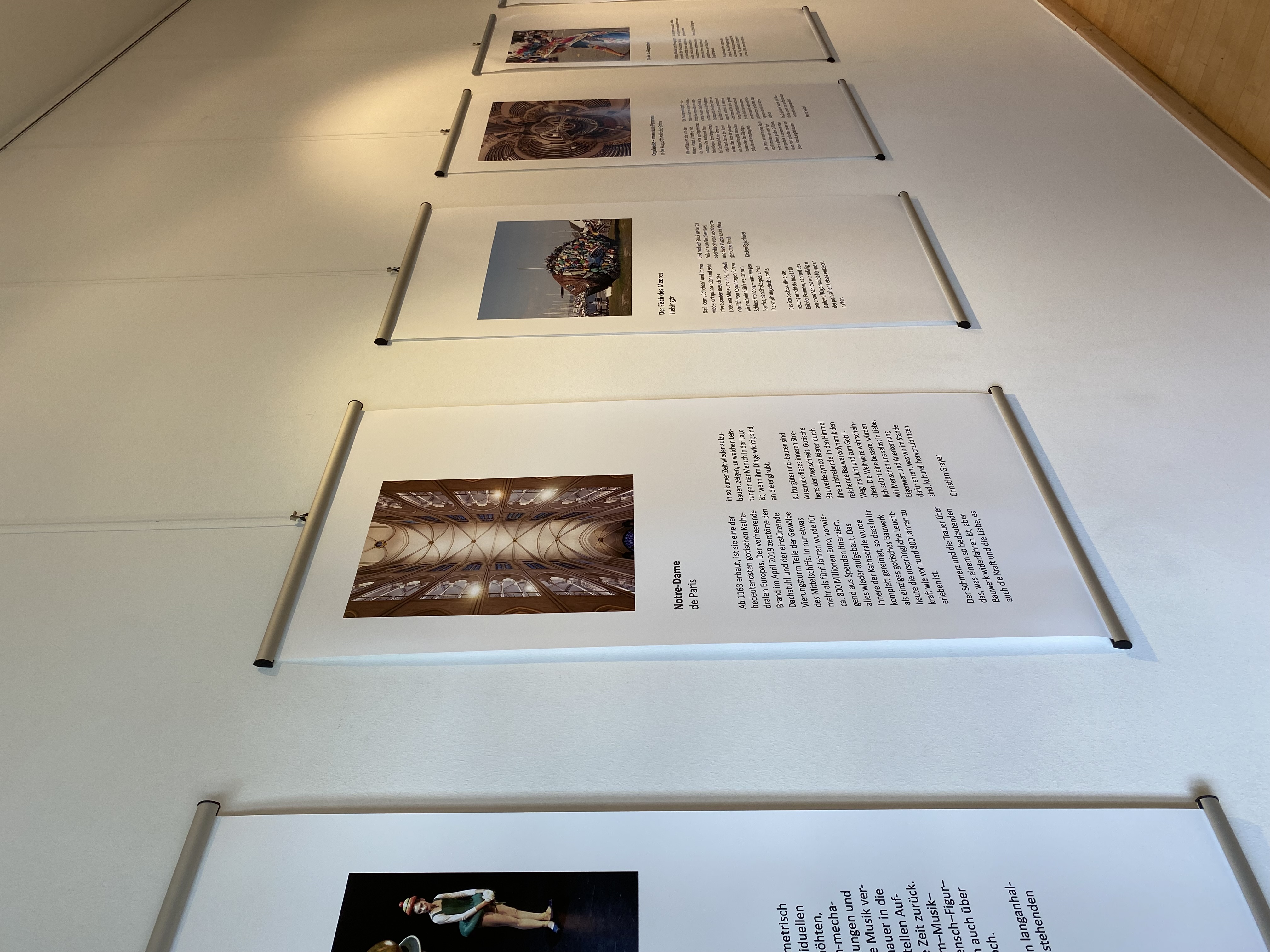 Mehrere großformatige, hochformatige Plakate hängen nebeneinander an einer weißen Wand. Jedes Plakat zeigt oben ein Farbfoto und darunter einen längeren Textabschnitt. Die Motive der Fotos sind unterschiedlich und reichen von Menschen über Architektur bis zu Gruppenaufnahmen. Die Plakate sind mit Metallleisten oben und unten befestigt und hängen an dünnen Drähten von der Decke. Der Boden ist hell und glatt.