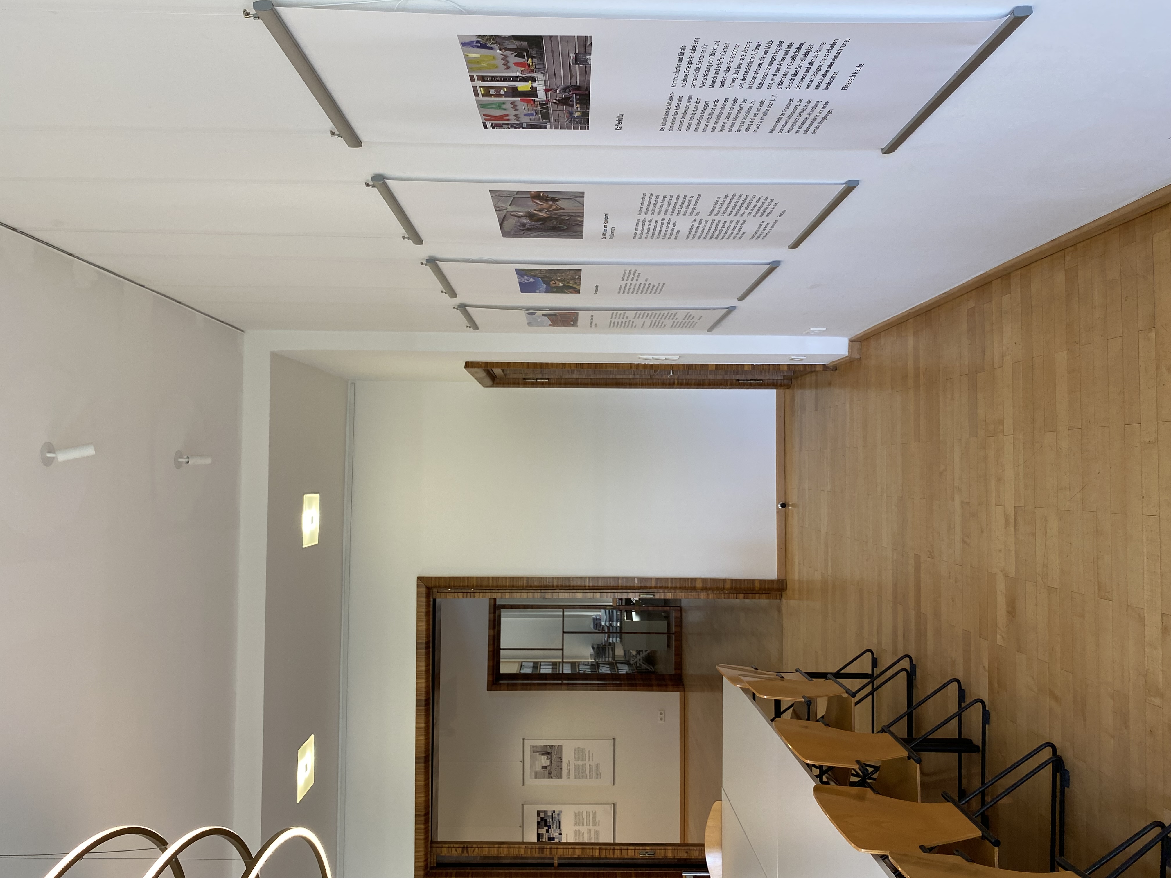 Heller Ausstellungsraum mit Holzfußboden und weißen Wänden. An der rechten Wand hängen mehrere großformatige Tafeln mit Text und Bildern, die in Metallrahmen befestigt sind. Links steht ein langer Tisch mit hellen Stühlen. Im Hintergrund sind weitere Räume mit ähnlicher Gestaltung durch offene Türrahmen sichtbar.