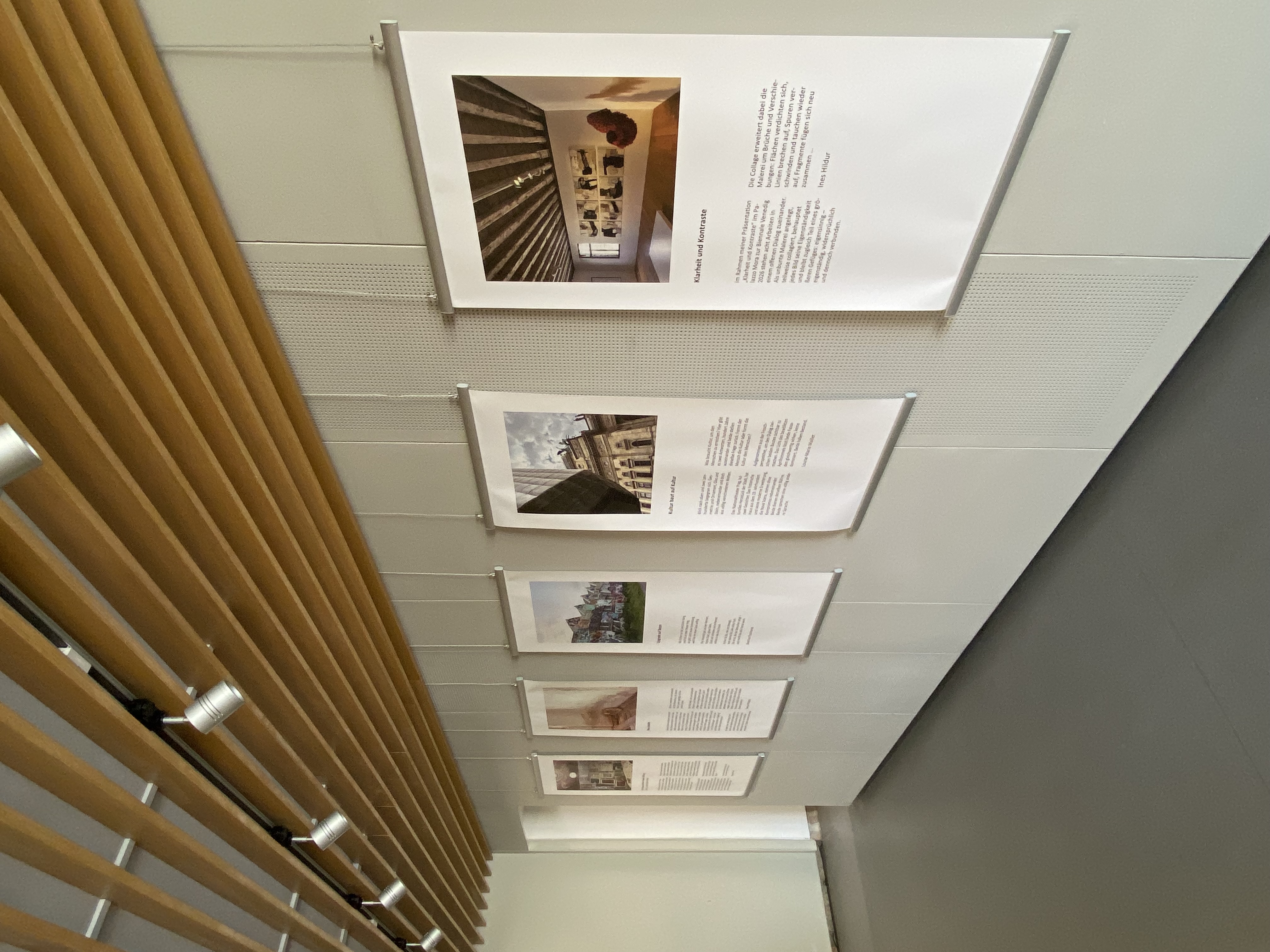 Mehrere großformatige Plakate hängen nebeneinander an einer hellen Wand. Jedes Plakat zeigt ein Foto mit einem längeren Textabschnitt darunter. Die Motive der Fotos variieren und zeigen Innen- und Außenansichten von Gebäuden. Die Plakate sind mit Drahtseilen an einer Holzdecke befestigt, deren Lamellen parallel verlaufen. Der Boden ist grau und glatt, der Raum wirkt modern und aufgeräumt.