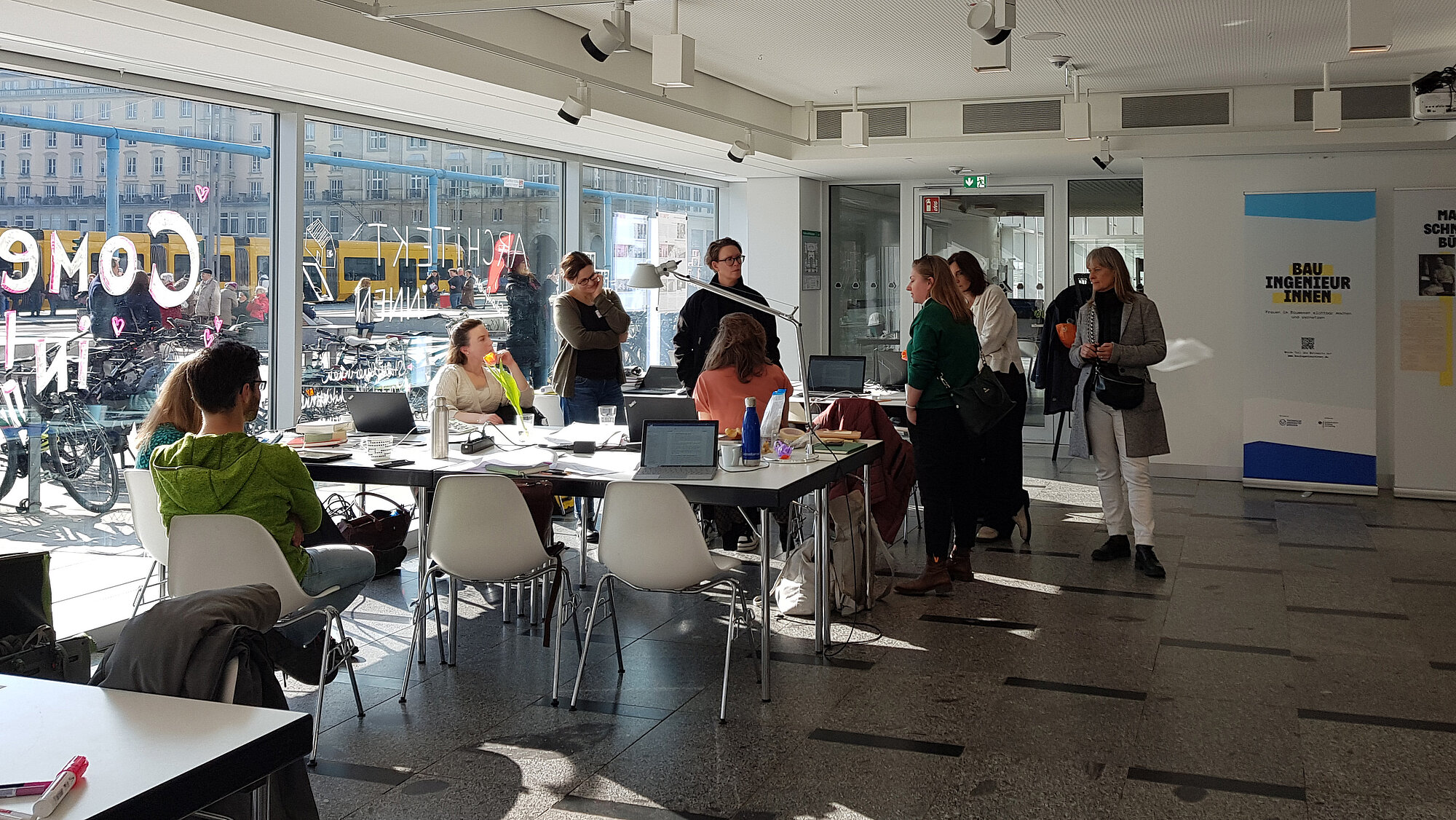 eine Gruppe von Frauen, sitzend und stehend, um Schreibtische und Stühle versammelt und in verbalem Austausch. Die linke Seite des Raumes ist von raumgroßen Glasscheiben flankiert, die das Stadtleben draußen zeigen.