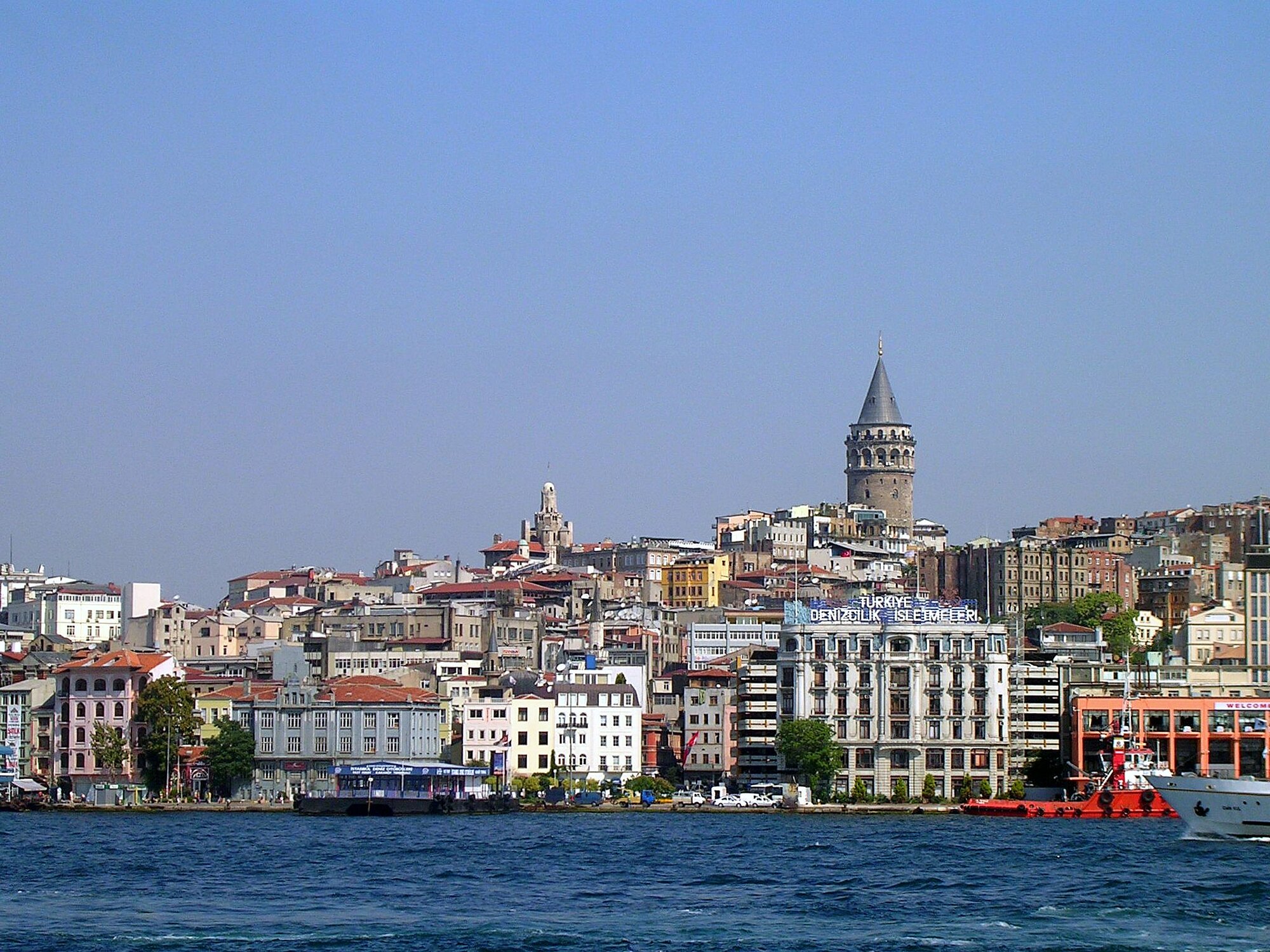 Blick auf Istanbul (Lars-Arne Dannenberg) Stadtansicht mit dicht bebauten, mehrstöckigen Gebäuden am Ufer eines breiten Gewässers. Im Hintergrund ragt ein hoher, runder Turm mit spitzem Dach über die umliegenden Häuser hinaus. Die Fassade der Gebäude zeigt unterschiedliche Farben und Baustile. Im Vordergrund ist das Wasser ruhig, am Ufer liegen mehrere Boote und Schiffe. Der Himmel ist wolkenlos und blau.