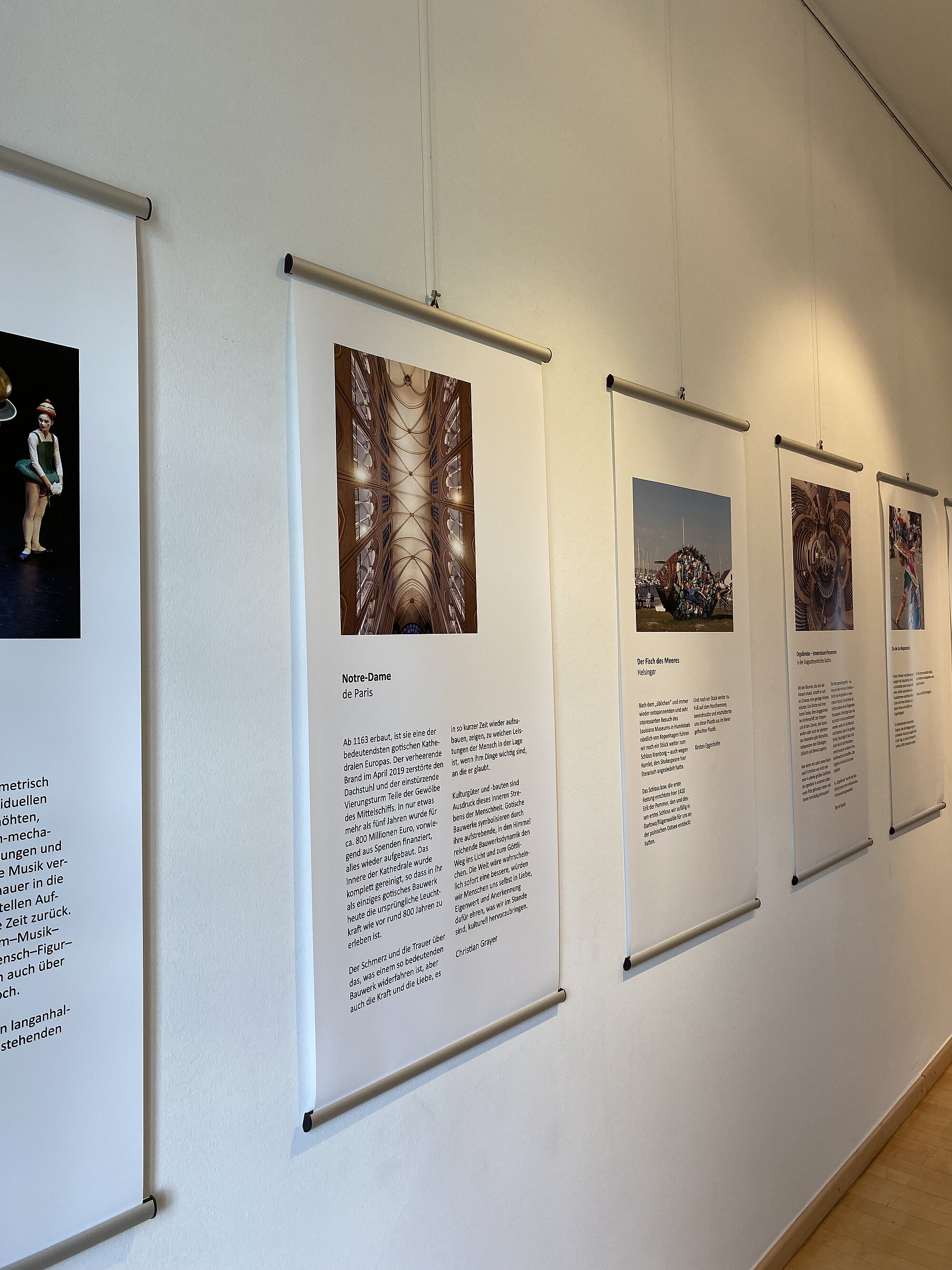(Architektenkammer Sachsen) Mehrere großformatige, hochformatige Plakate hängen nebeneinander an einer weißen Wand. Jedes Plakat zeigt oben ein Farbfoto und darunter einen längeren Textabschnitt. Die Motive der Fotos sind unterschiedlich und reichen von Menschen über Architektur bis zu Gruppenaufnahmen. Die Plakate sind mit Metallleisten oben und unten befestigt und hängen an dünnen Drähten von der Decke. Der Boden ist hell und glatt.