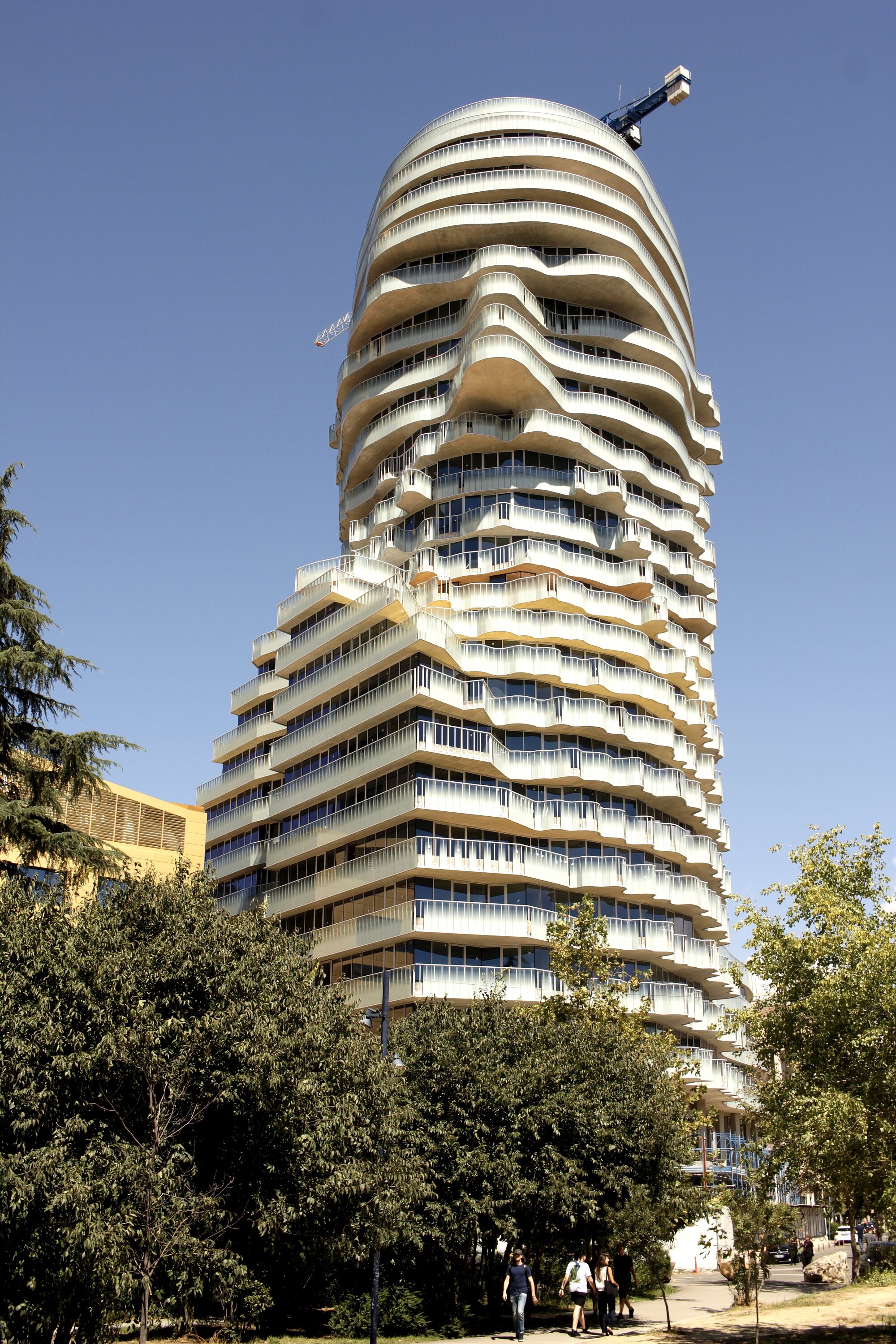 Tirana's Rock oder Skanderberg Building in Tirana (Von Tom Ackroyd - Eigenes Werk, CC BY-SA 4.0, https://commons.wikimedia.org/w/index.php?curid=175780876) Ein modernes Hochhaus mit wellenförmig geschwungenen, übereinander gestapelten Etagen dominiert die Szene. Die Fassade besteht aus hellen, horizontal verlaufenden Elementen und zahlreichen Balkonen. Auf dem Dach befindet sich ein Baukran. Im Vordergrund stehen Bäume, die einen Teil des Gebäudes verdecken. Der Himmel ist wolkenlos und blau.