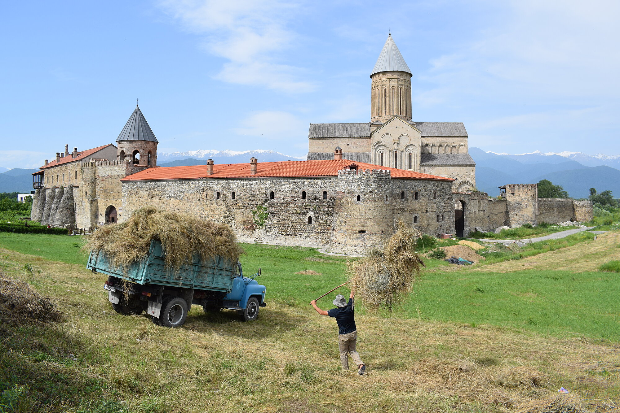 Alawerdi Kathedrale in der Region Kachetien (Dr. Lars-Arne Dannenberg) Im Vordergrund lädt eine Person mit erhobenen Armen Heu von einem Feld auf einen blauen Lastwagen. Der Lastwagen steht seitlich am Feldrand, die Ladefläche ist bereits mit Heu beladen. Im Hintergrund erhebt sich eine große Klosteranlage mit dicken Steinmauern, mehreren Türmen und einem hohen Kirchenschiff mit Kuppel. Die Dächer sind ziegelrot gedeckt. Dahinter sind schneebedeckte Berge zu sehen, der Himmel ist klar.